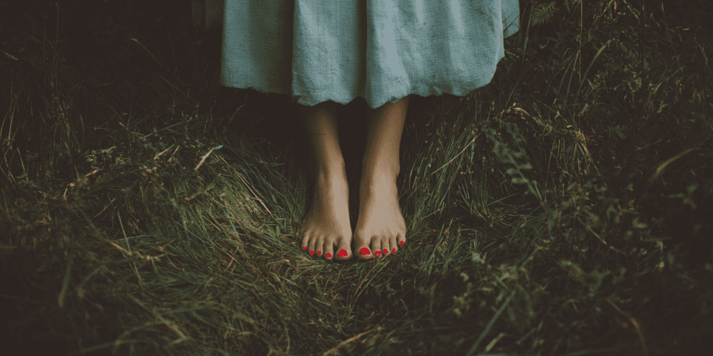 Bare feminine feet standing barefoot on lush green forest floor with small wildflowers and grass, natural soft light filtering through trees, grounding in nature, ancestral living style